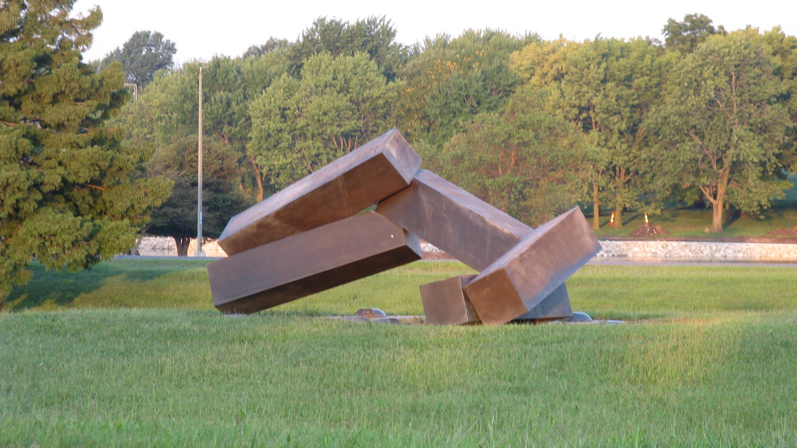 Rust-colored abstract metal sculpture of interlocking beams resting on grass, with trees and a stone wall in the background