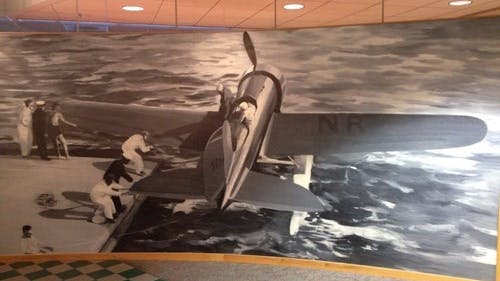Black-and-white mural of a vintage airplane on a runway surrounded by people, with a stylized ocean in the background.
