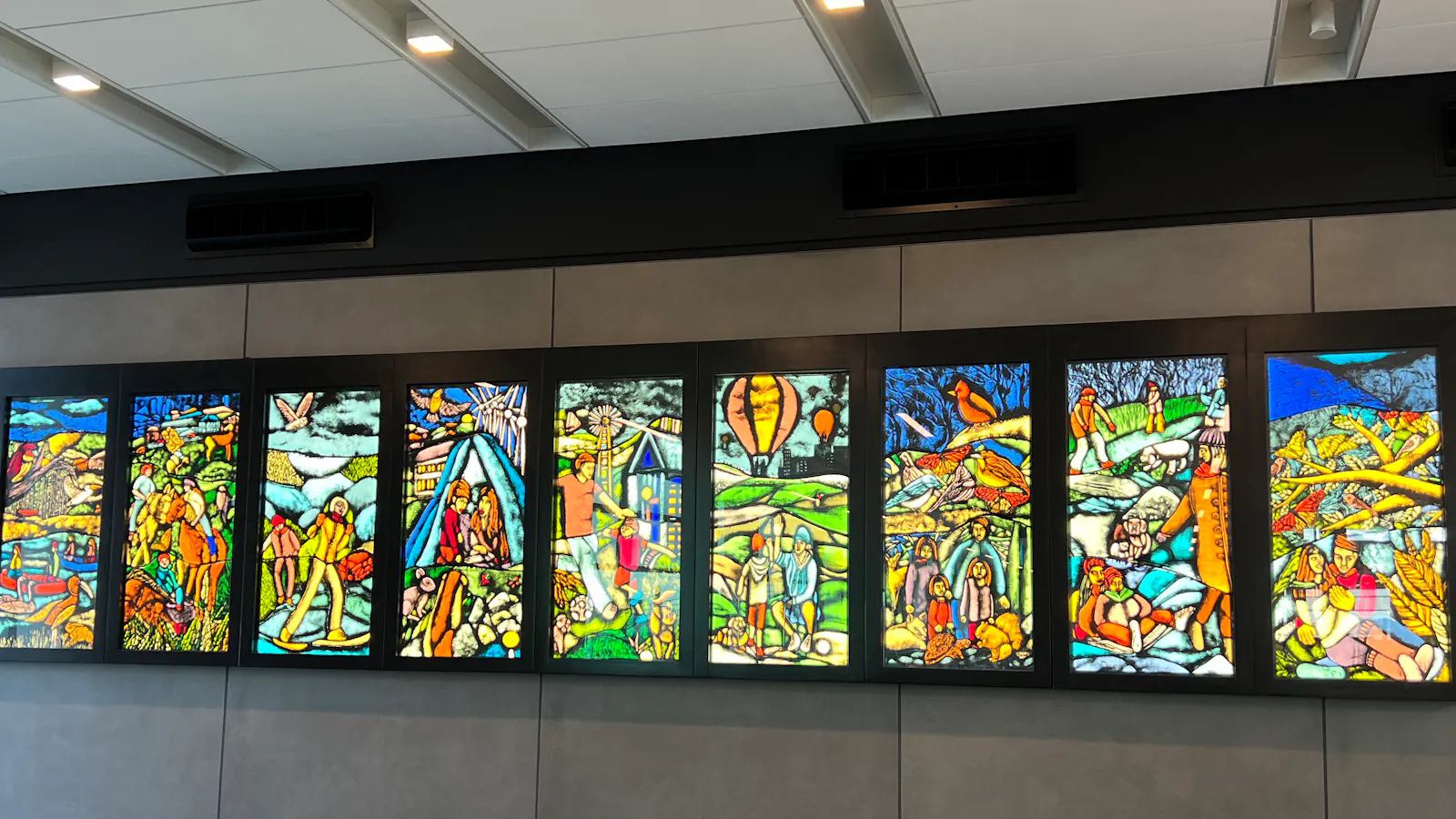 Large mural composed of ten vertical panels resembling stained glass, each depicting vibrant, storybook-like scenes of people, landscapes, and community life in vivid color.