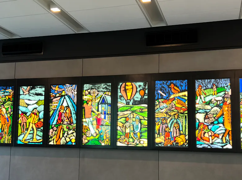 Large mural composed of ten vertical panels resembling stained glass, each depicting vibrant, storybook-like scenes of people, landscapes, and community life in vivid color.