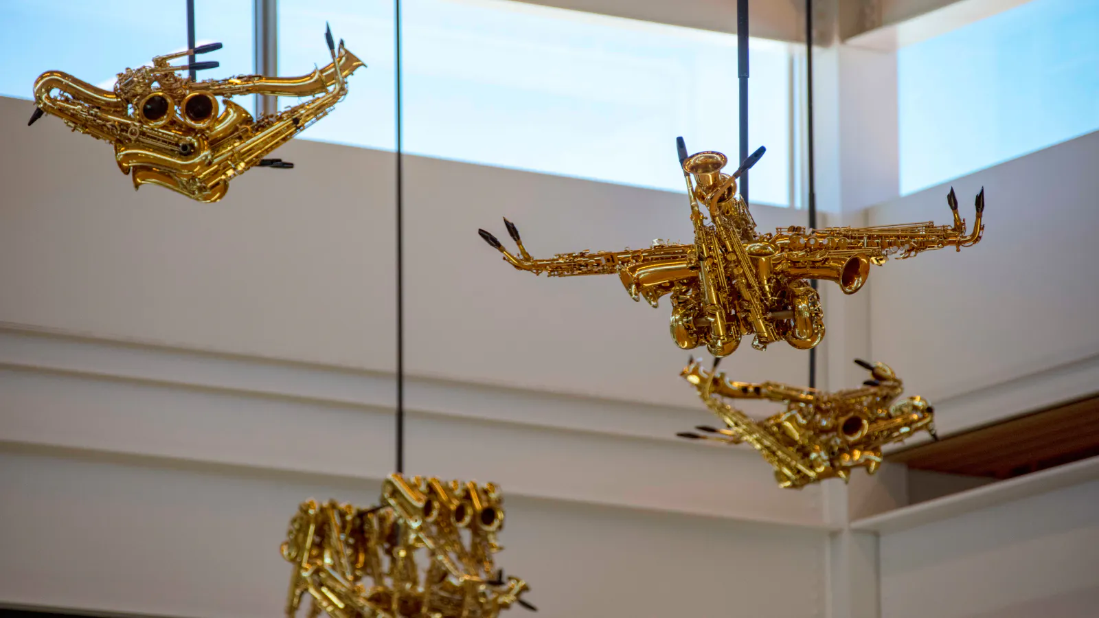 Four golden sculptures made from saxophones and other brass instruments hang from the ceiling in an airport terminal. Each piece is shaped like a stylized bird or aircraft, suspended by black cords against a backdrop of white walls and high windows.