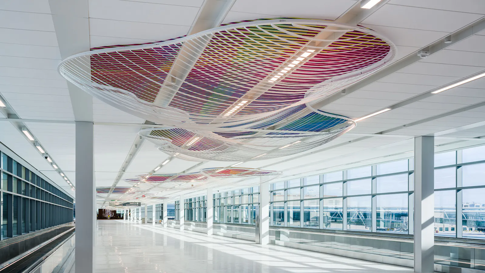 Ceiling installation in a bright airport corridor featuring a series of large, wavy white mesh forms filled with multicolored lattice patterns that cast dynamic shadows on the floor below.
