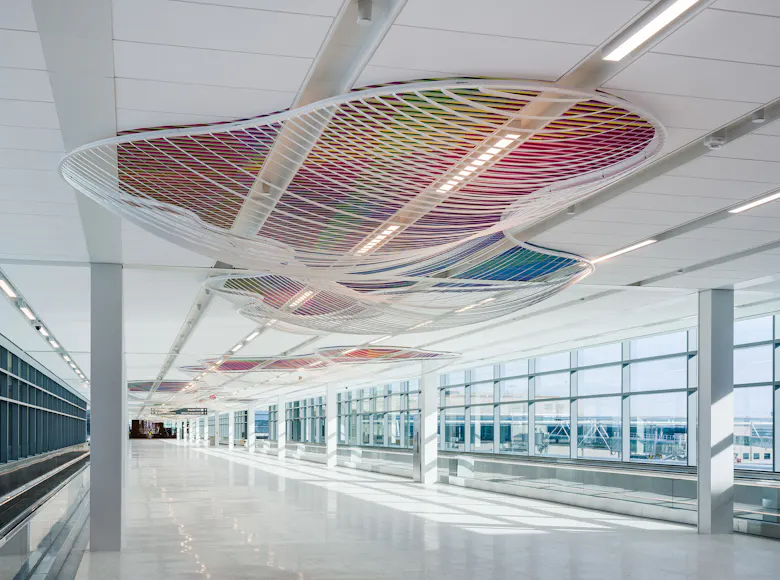 Ceiling installation in a bright airport corridor featuring a series of large, wavy white mesh forms filled with multicolored lattice patterns that cast dynamic shadows on the floor below.
