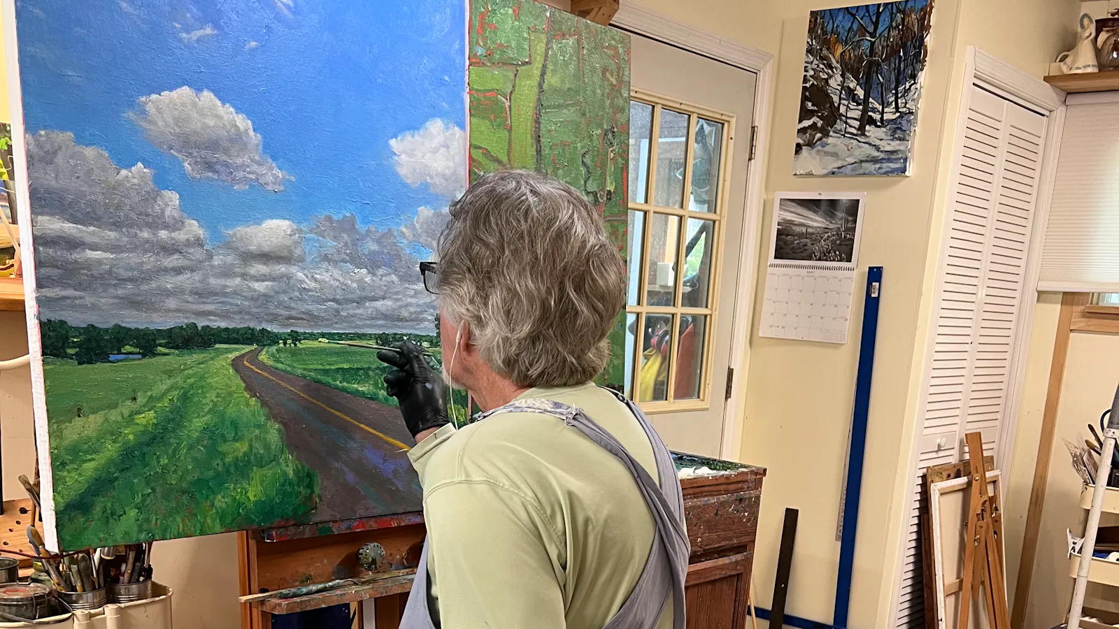 Artist John Louder seated in a studio painting a rural road landscape under a bright sky with clouds, referencing a map texture on the right side of the canvas.