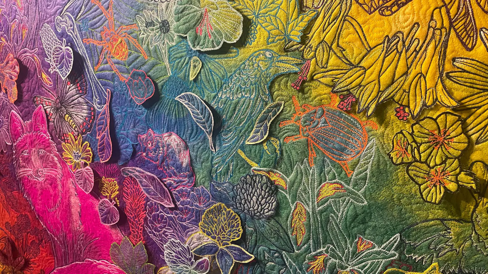 Detailed textile art showing a rainbow-colored forest scene with a pink fox, butterflies, beetles, birds, and dense embroidered foliage.