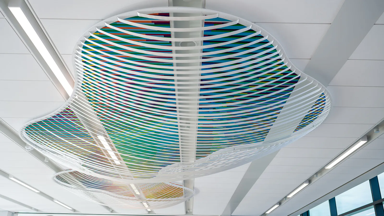 Colorful ceiling sculpture with undulating white lattice structure and diagonal rainbow bands suspended above a wide airport walkway with floor-to-ceiling windows and moving sidewalks.