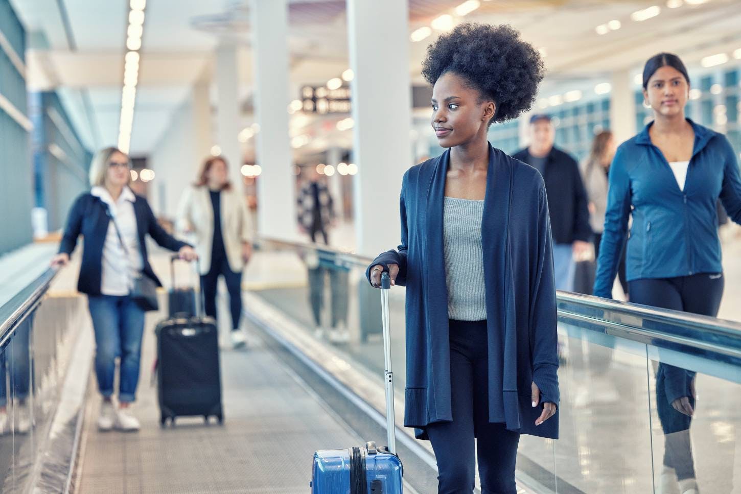 Glass-walled airport concourse with moving walkways and skylights, showing travelers walking through.