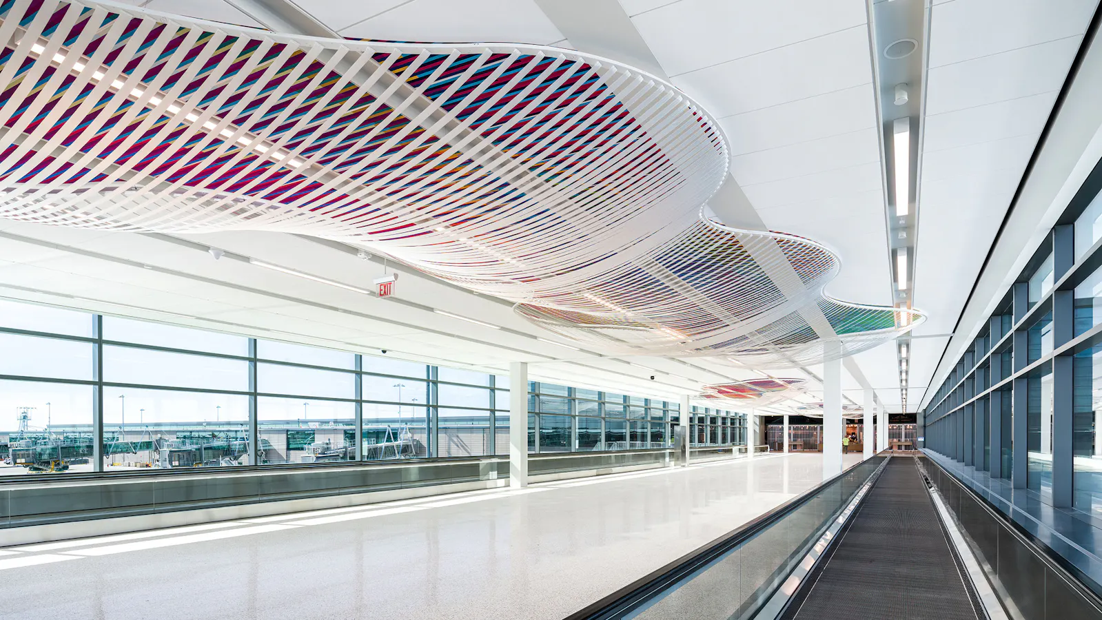 Perspective view of a moving walkway beneath a flowing ceiling installation made of white sculptural frames filled with vibrant, interwoven ribbons of color, set against large terminal windows.