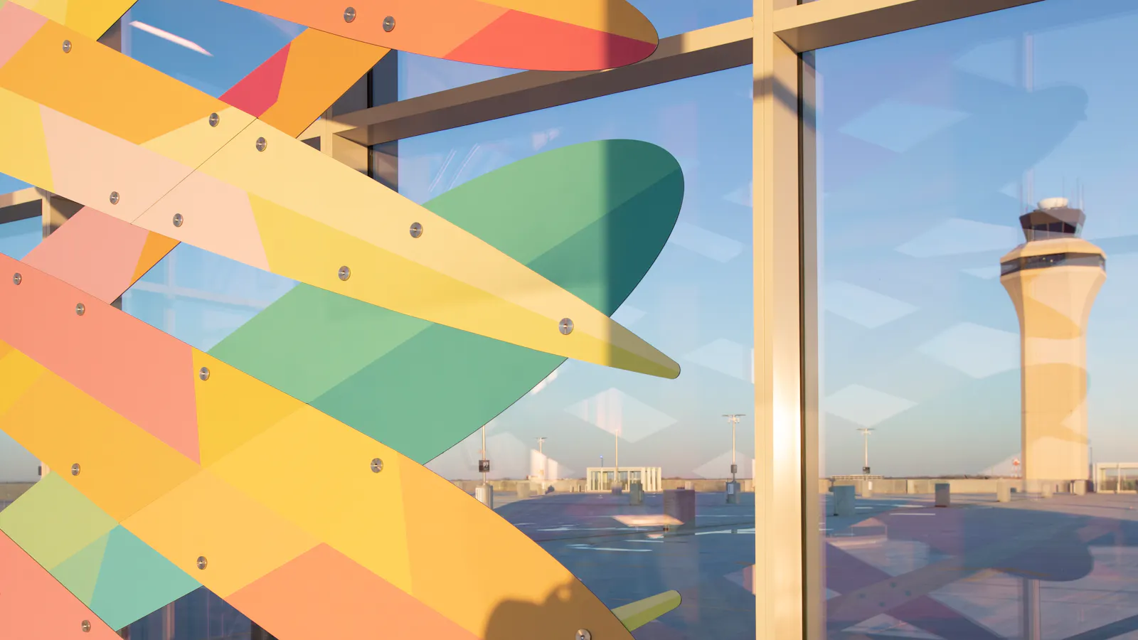 Close-up of a colorful layered sculpture inside Kansas City International Airport's parking garage, with large windows showing the airfield and control tower in the background.