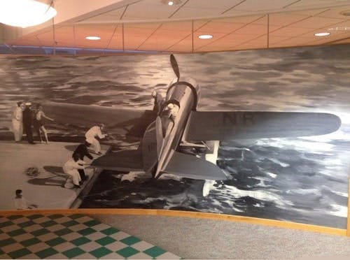 Black-and-white mural of a vintage airplane on a runway surrounded by people, with a stylized ocean in the background.