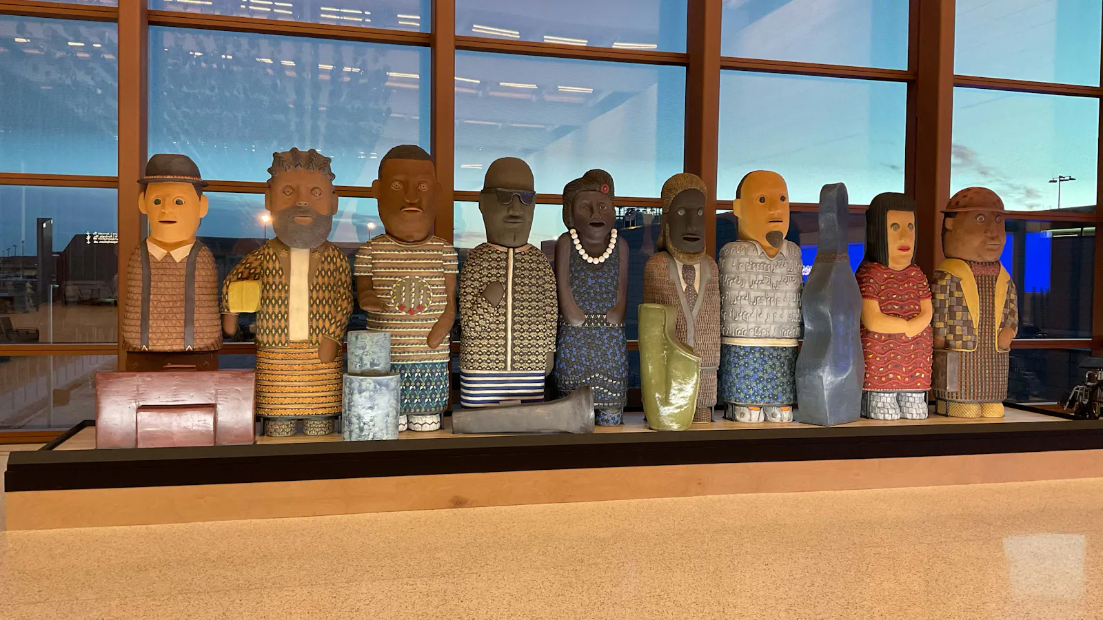 A row of eleven colorful, stylized human sculptures, each uniquely patterned and posed, displayed indoors at Kansas City International Airport.