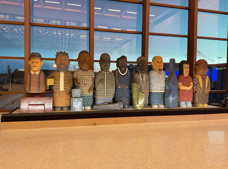 A row of eleven colorful, stylized human sculptures, each uniquely patterned and posed, displayed indoors at Kansas City International Airport.