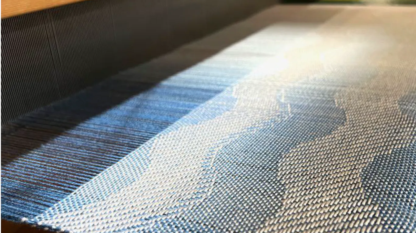 Close-up of blue and white woven fabric on a loom, featuring a wave-like pattern and subtle gradient transitioning from dark to light tones.
