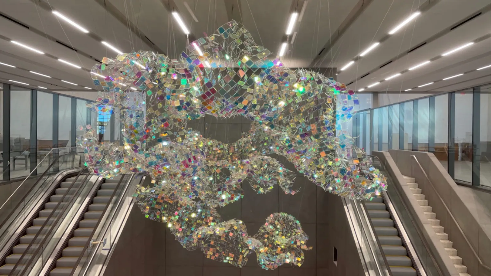 Suspended sculpture of iridescent, net-like mesh glimmering above escalators at Kansas City International Airport.