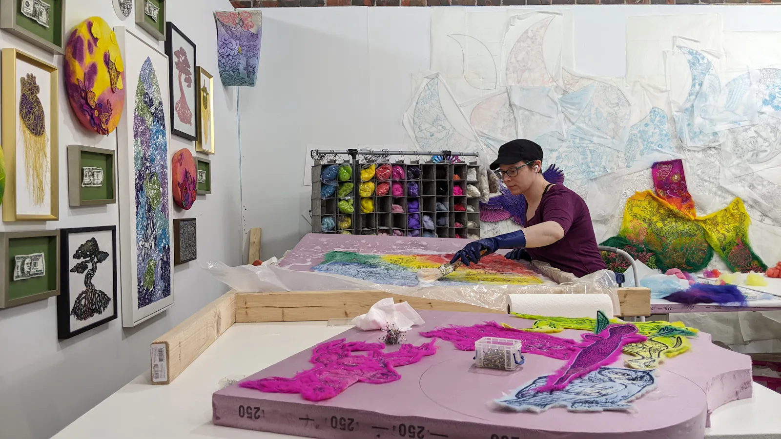 Artist Rachelle Gardner-Roe working in a studio on a large textile piece with rainbow colors, surrounded by wool materials, pinned fabric, and framed fiber art on the wall.