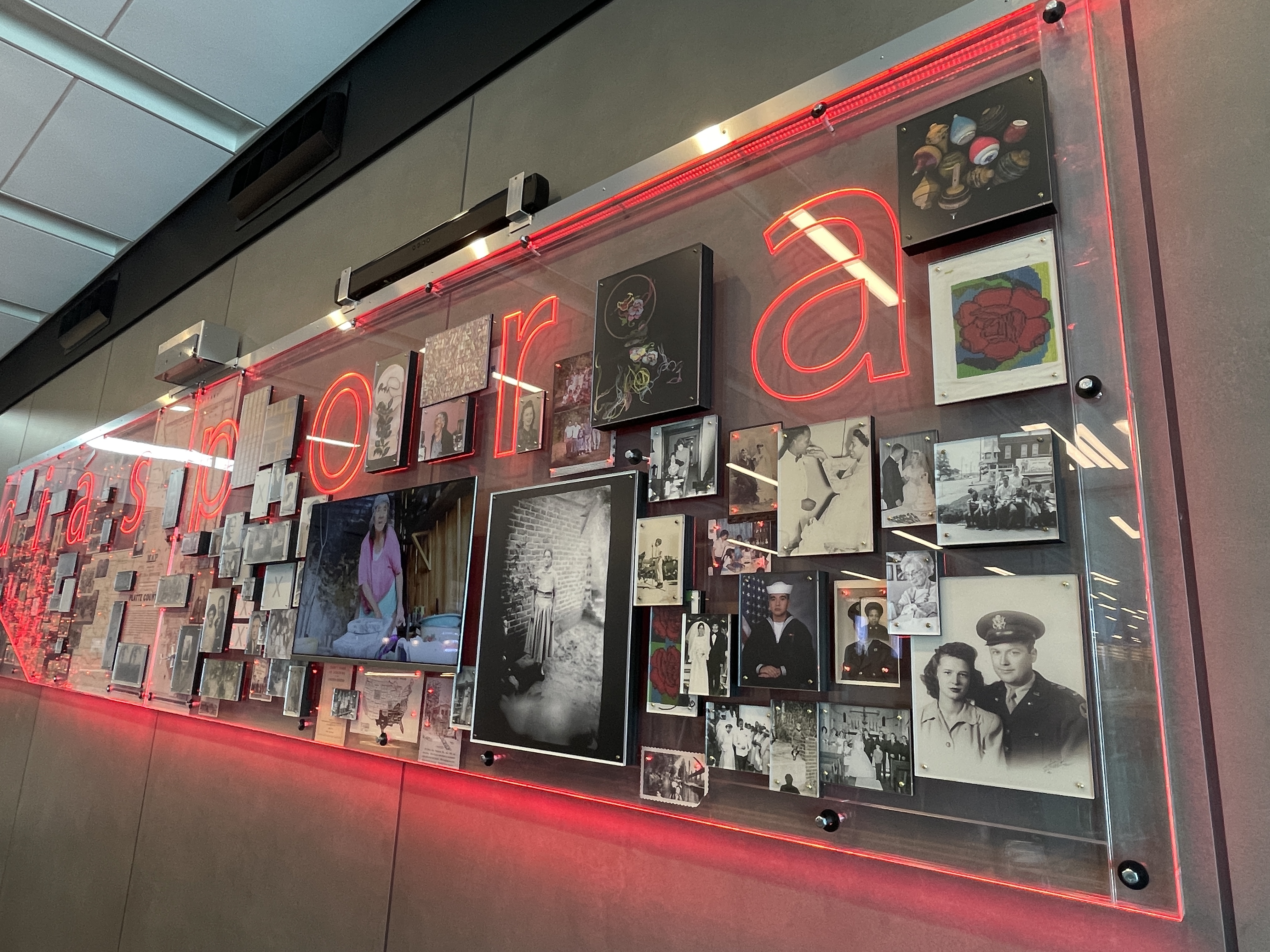 Angled view of the “diasporia” installation’s right end, showcasing family portraits, military photos, and embroidered artworks arranged under red neon lighting on a glass panel.