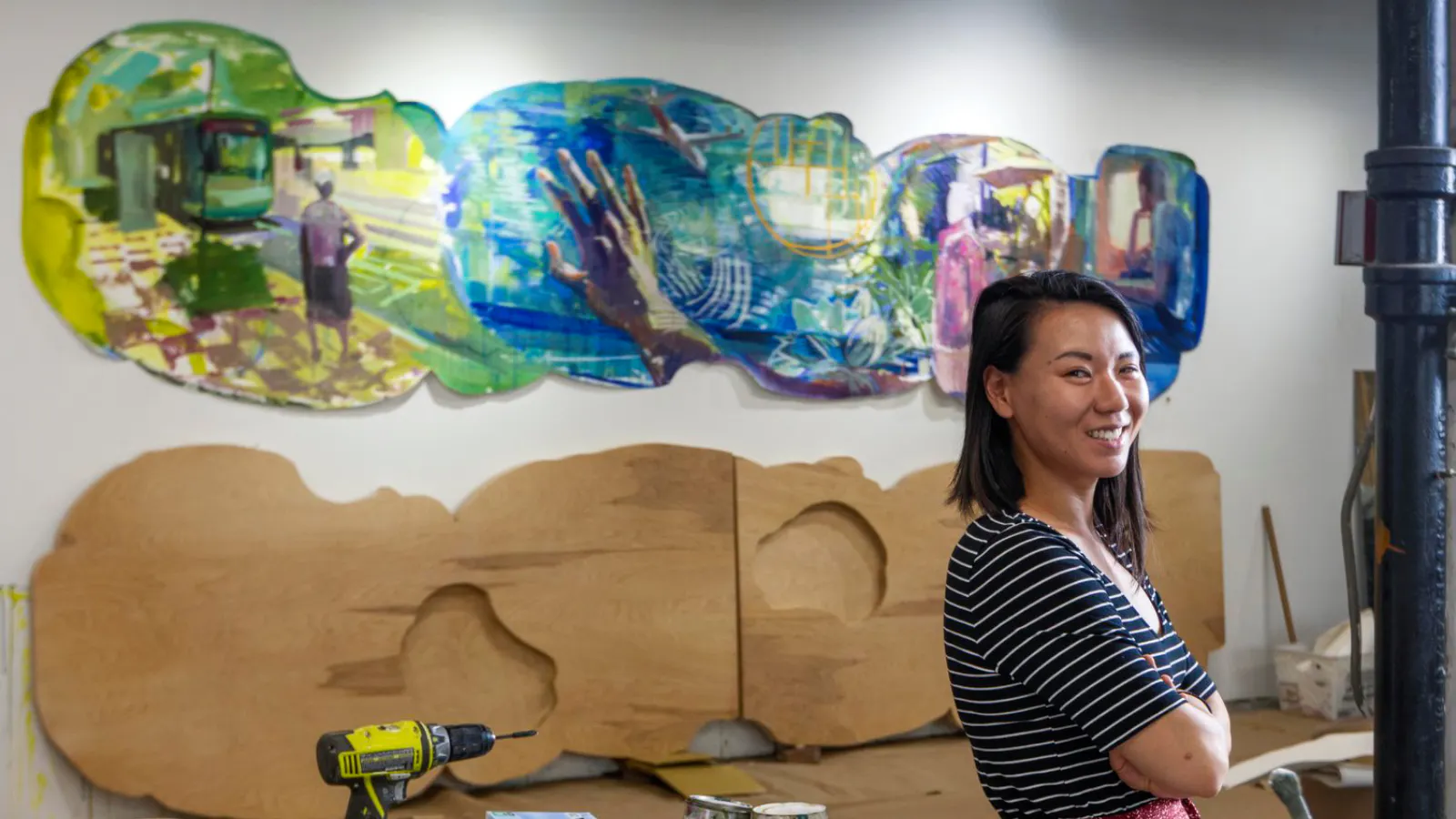 Artist Kathy Liao standing in front of a colorful memory-themed mural in progress, featuring layered scenes and wooden backing panels.