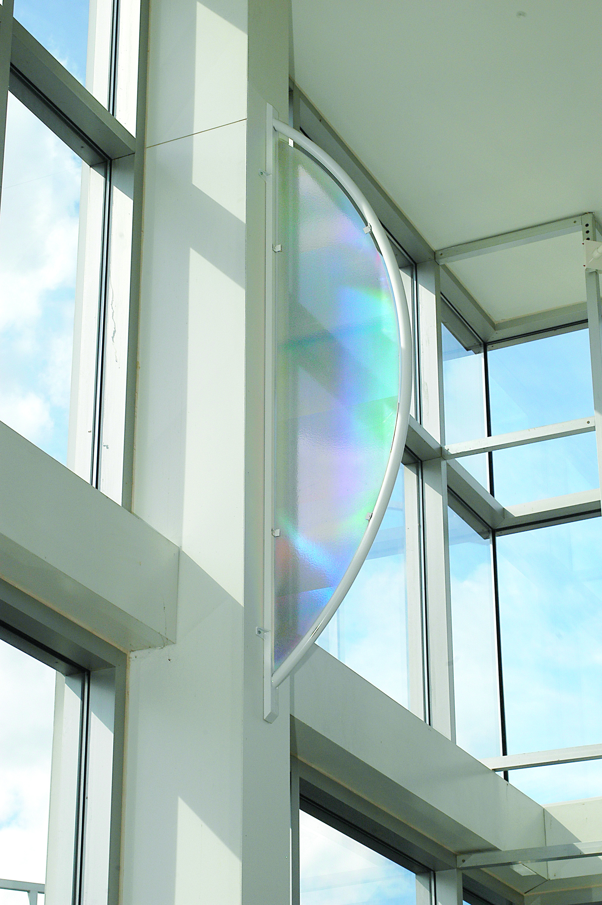 Translucent, iridescent panel in a curved white frame mounted on a column inside a glass-walled atrium, reflecting soft rainbow colors from natural light.