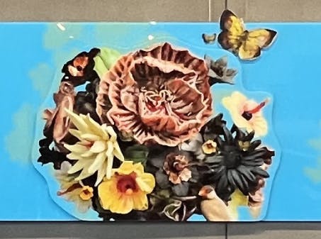 Colorful mural on a bright blue background featuring blooming flowers, butterflies, and a surprised woman’s face, blending realism with collage-like elements.
