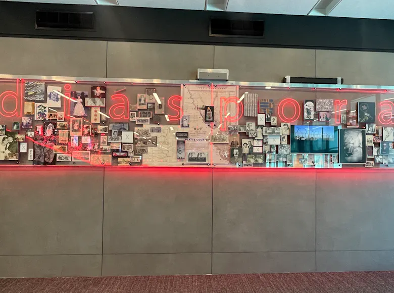Wall-mounted art installation featuring a collage of historical photos, documents, and small artifacts arranged behind glass with the word “diasporia” illuminated in red neon.