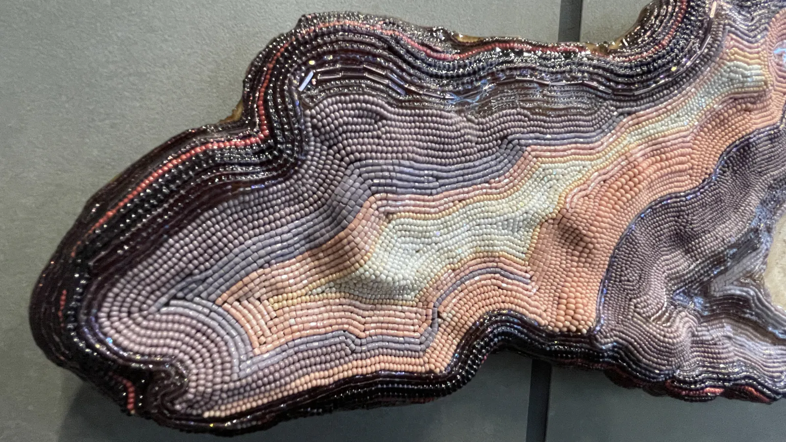 Extreme close-up of a sculptural edge composed of tightly arranged beads in layered, undulating rows of purple, pink, cream, and black, mimicking geological strata.
