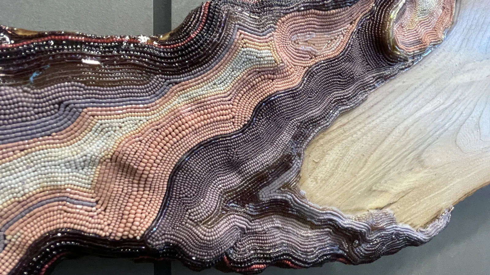 Detail of a beaded wood sculpture showing a seamless transition from smooth natural wood to densely packed, wavy rows of beads in shades of mauve, peach, lavender, and black.
