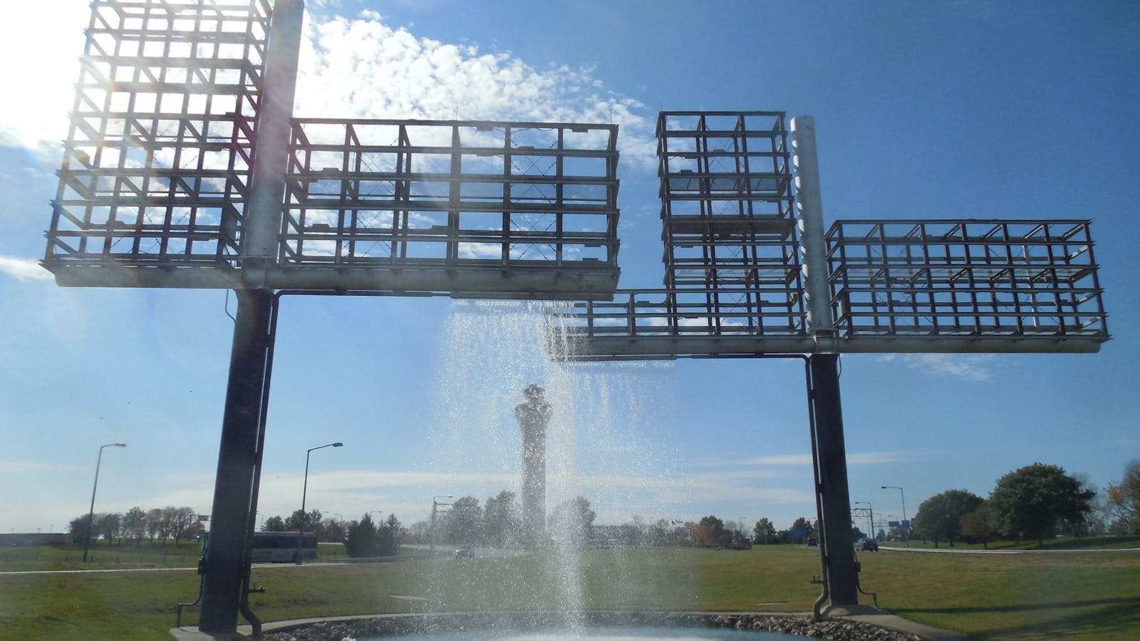 Daylight view of a tall metal grid fountain structure with water falling into a circular pool, and the airport control tower visible in the distance.