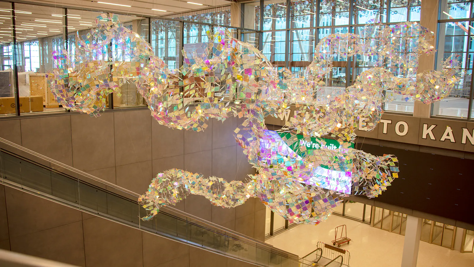 Flowing, iridescent mesh sculpture suspended over an escalator near “Welcome to Kansas City” signage at Kansas City International Airport.