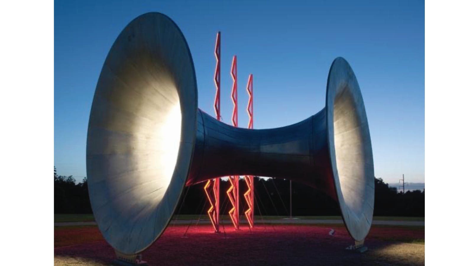 Large, spotlighted metal sculpture resembling a spool or horn with red zigzag columns in the background, set against a twilight sky.