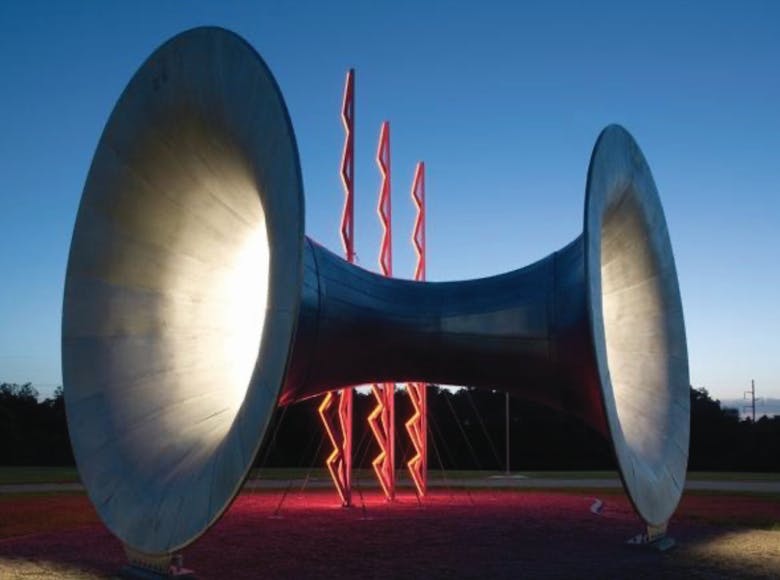 Large, spotlighted metal sculpture resembling a spool or horn with red zigzag columns in the background, set against a twilight sky.