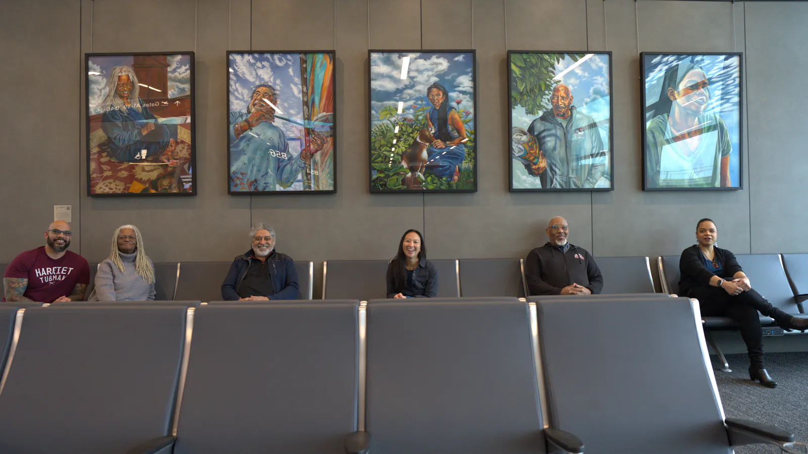 Six people sitting in airport seating beneath five portrait paintings on the wall at Kansas City International Airport, with each person matching the subject of a painting above them.