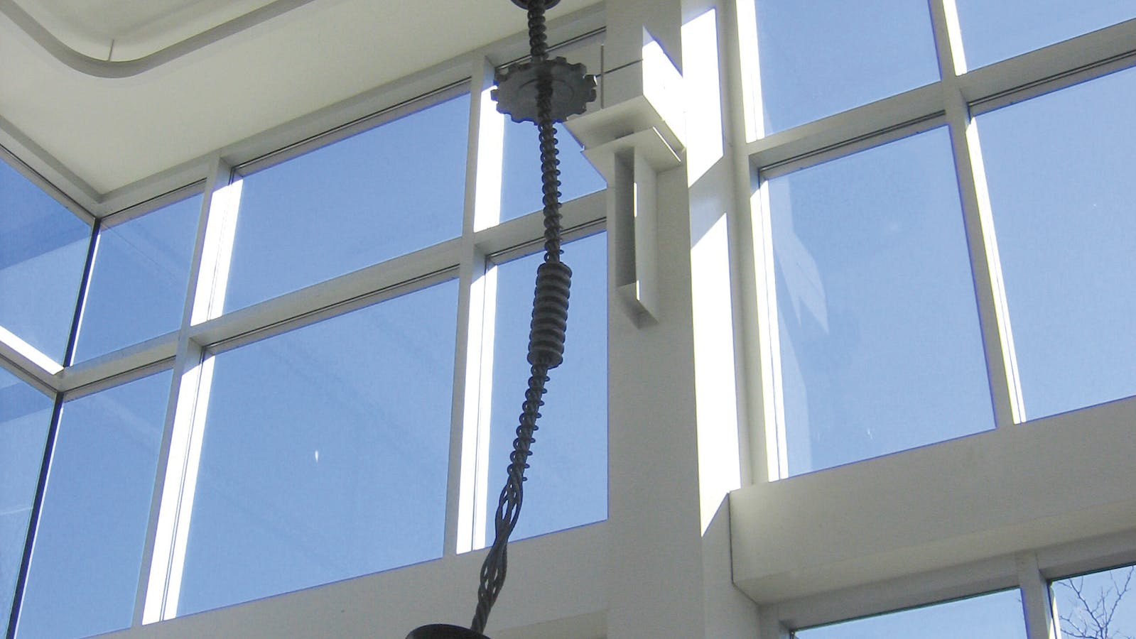 Ceiling-mounted sculpture featuring twisted black cables, metal coils, and spherical weights suspended in a glass-walled space with clear blue sky outside.