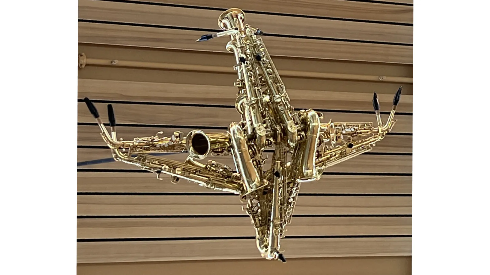 Sculpture resembling a bird or airplane, constructed entirely from brass instruments such as saxophones and trumpets, suspended from the ceiling.