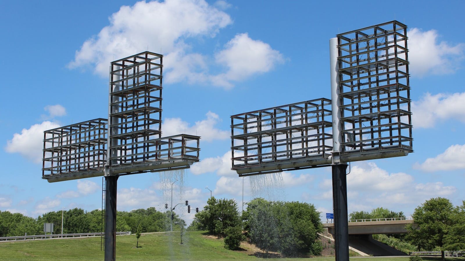 Daytime view of twin metal grid structures with water streaming down into a grassy median near a highway overpass.