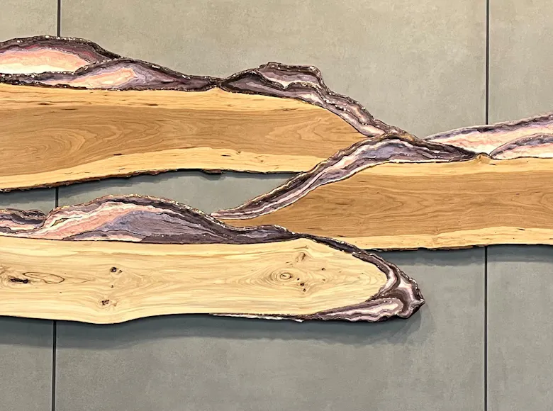 Wall-mounted sculpture composed of three elongated, organic slabs with live edges and layered purple and natural wood tones, evoking the form of windswept clouds or rolling landscapes.