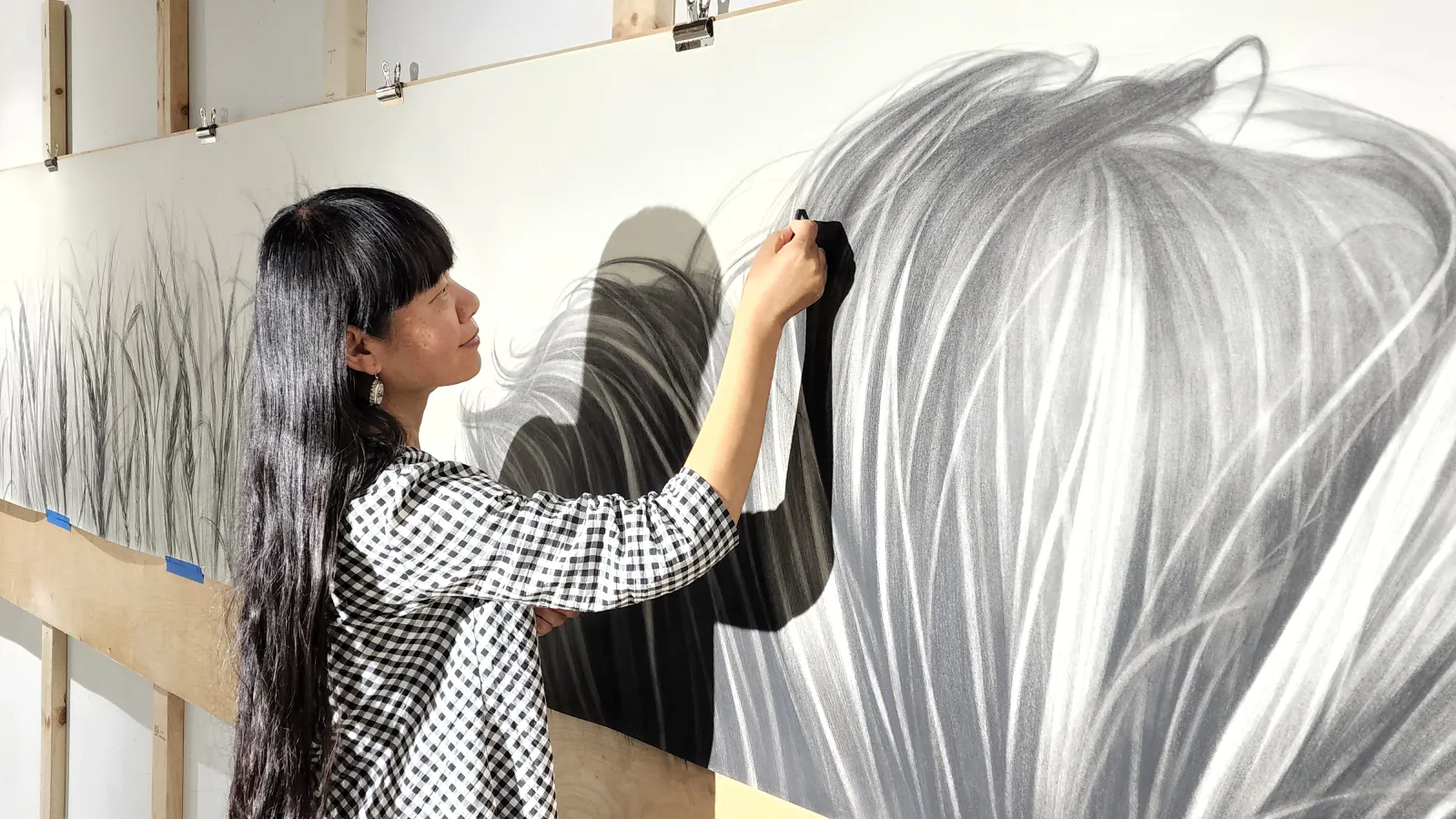 Artist Hong Chun Zhang working on a large charcoal drawing of flowing grass, blending strokes by hand on a long paper panel mounted to a studio wall.