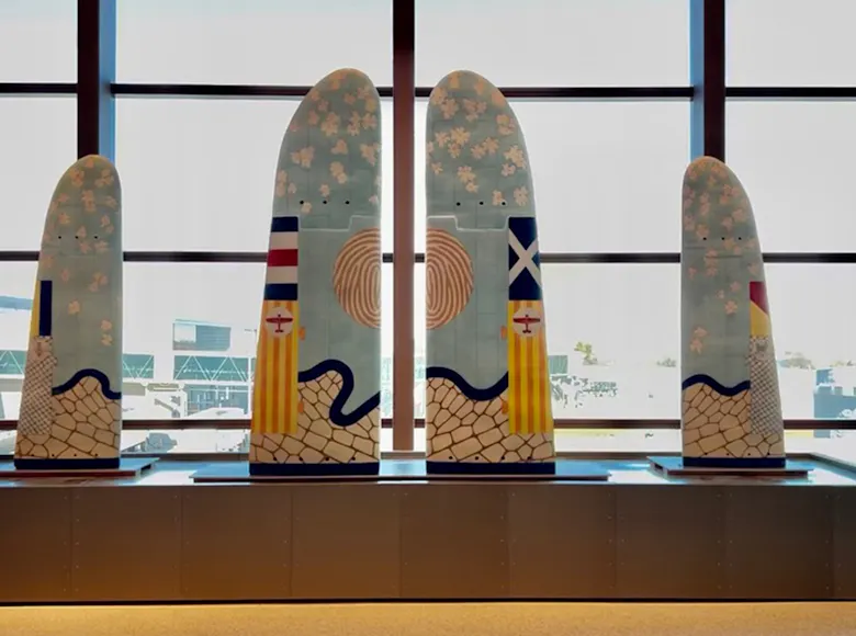 Five large, wing-shaped panels decorated with pastel patterns, abstract symbols, and flag-like designs stand upright in a row by a window at Kansas City International Airport.