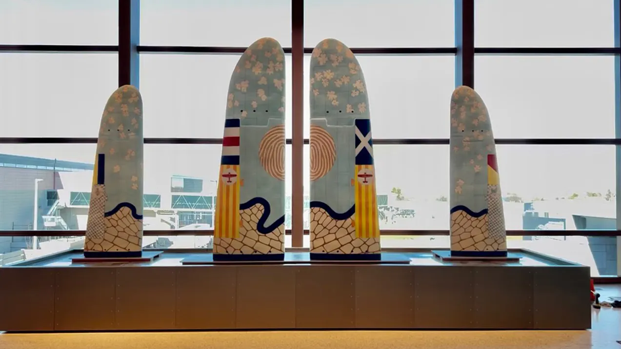 Five large, wing-shaped panels decorated with pastel patterns, abstract symbols, and flag-like designs stand upright in a row by a window at Kansas City International Airport.