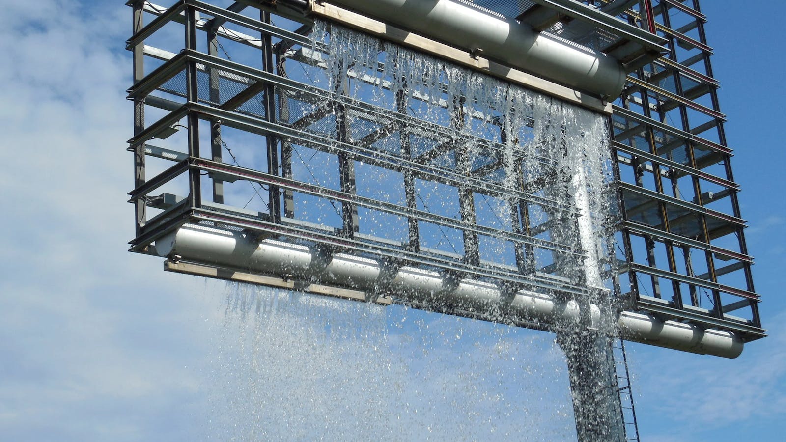 Close-up of cascading water flowing from a geometric metal fountain structure against a blue sky.