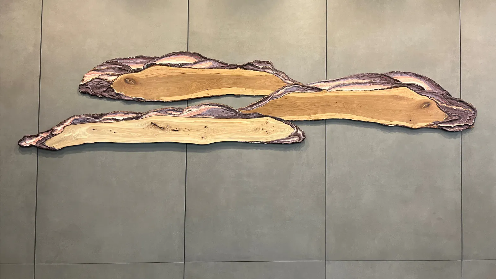 Wall-mounted sculpture composed of three elongated, organic slabs with live edges and layered purple and natural wood tones, evoking the form of windswept clouds or rolling landscapes.
