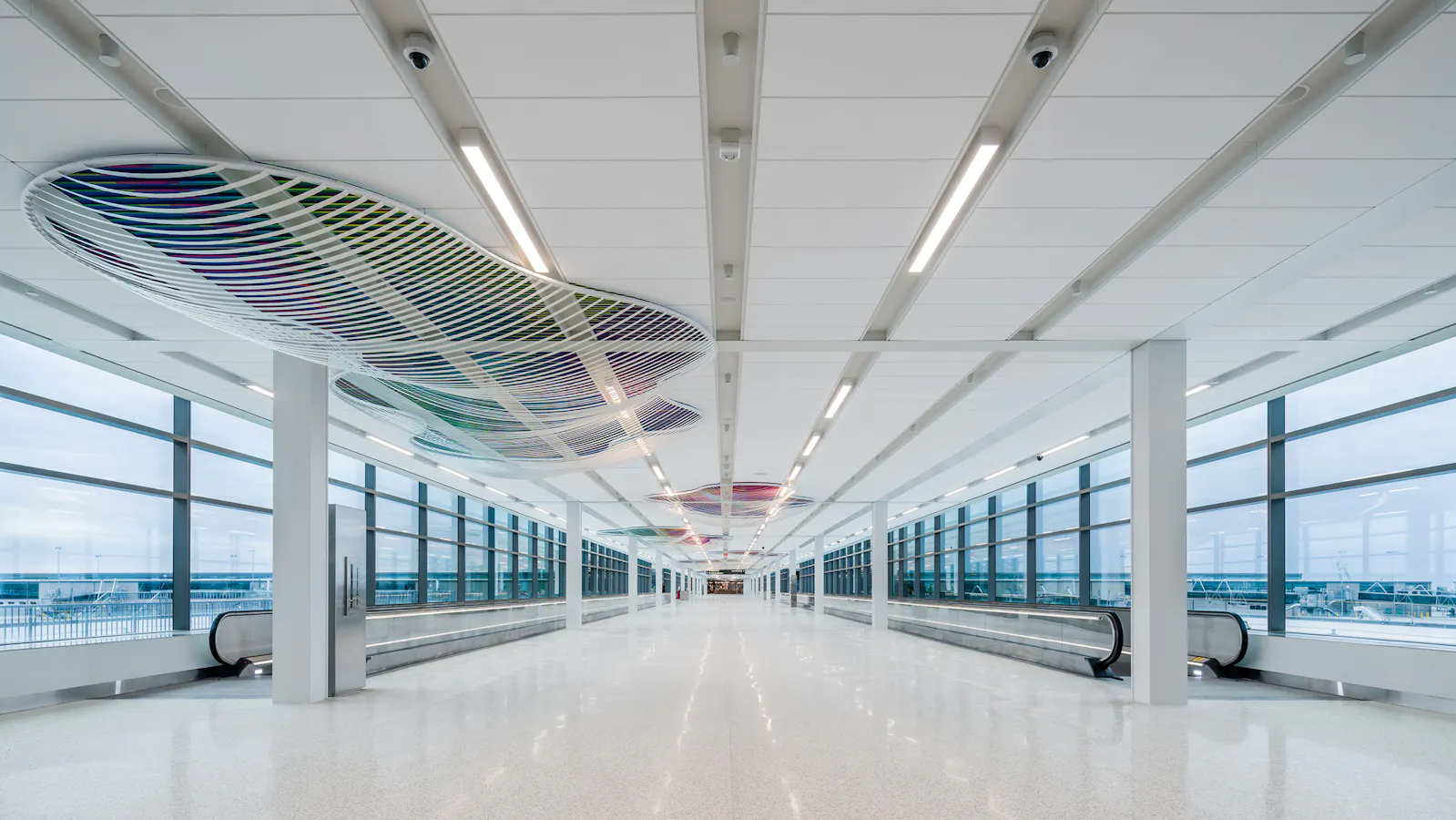Long airport corridor with ceiling-mounted sculptural forms made of white curved frames filled with colorful crisscrossed patterns, suspended above moving walkways and surrounded by large windows.