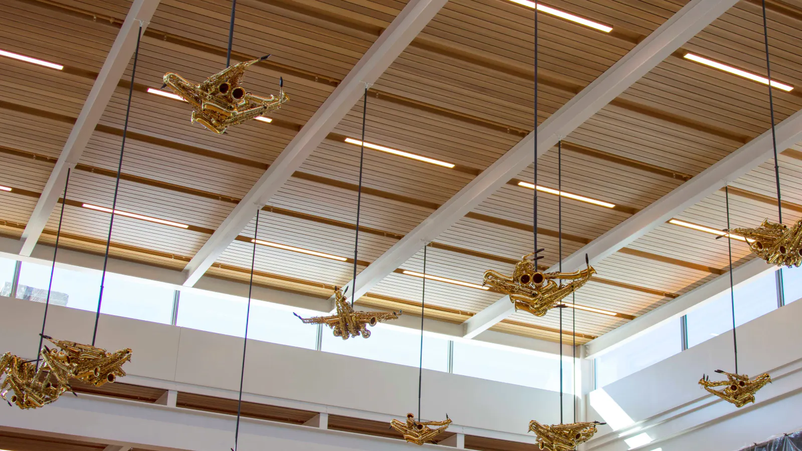 Sculptures resembling birds or airplanes made from golden brass instruments hang from the ceiling of an airport terminal. The pieces are suspended by black cables against a backdrop of wood-paneled ceiling beams and upper-level windows, allowing natural light to highlight the artwork.