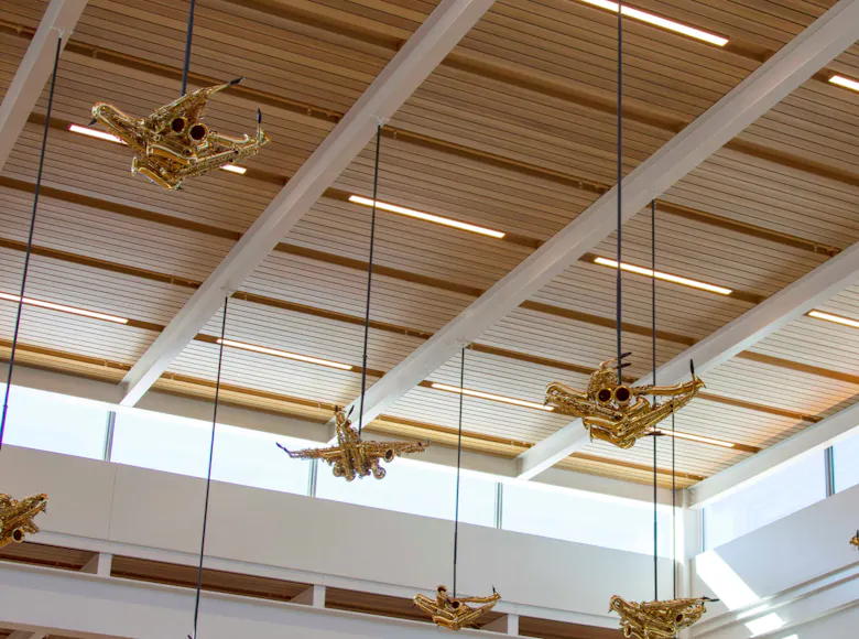 Sculptures resembling birds or airplanes made from golden brass instruments hang from the ceiling of an airport terminal. The pieces are suspended by black cables against a backdrop of wood-paneled ceiling beams and upper-level windows, allowing natural light to highlight the artwork.