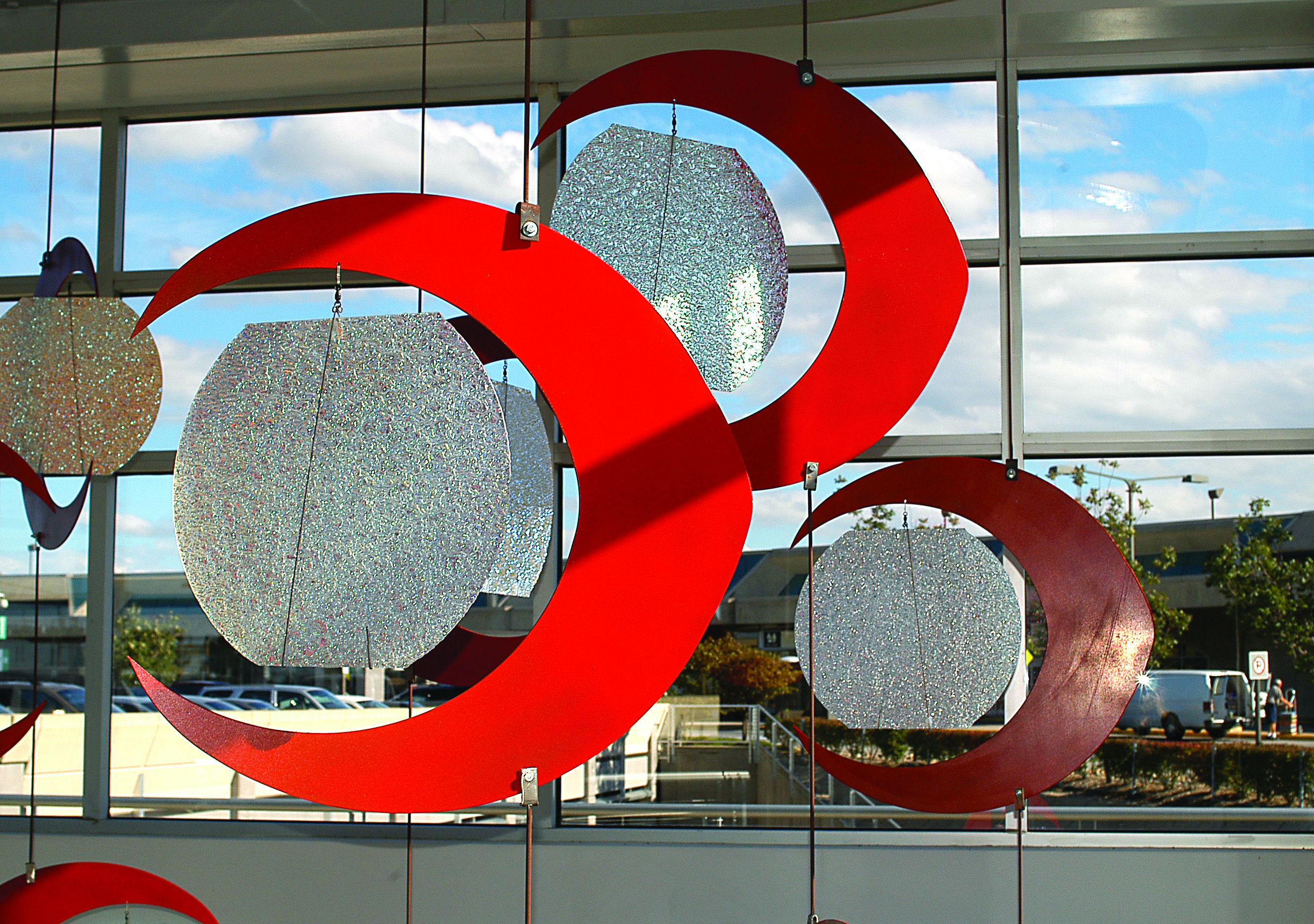 Close-up of a suspended mobile featuring glittery silver discs framed by bold red crescent shapes, set in front of large windows with a view of the airport exterior.