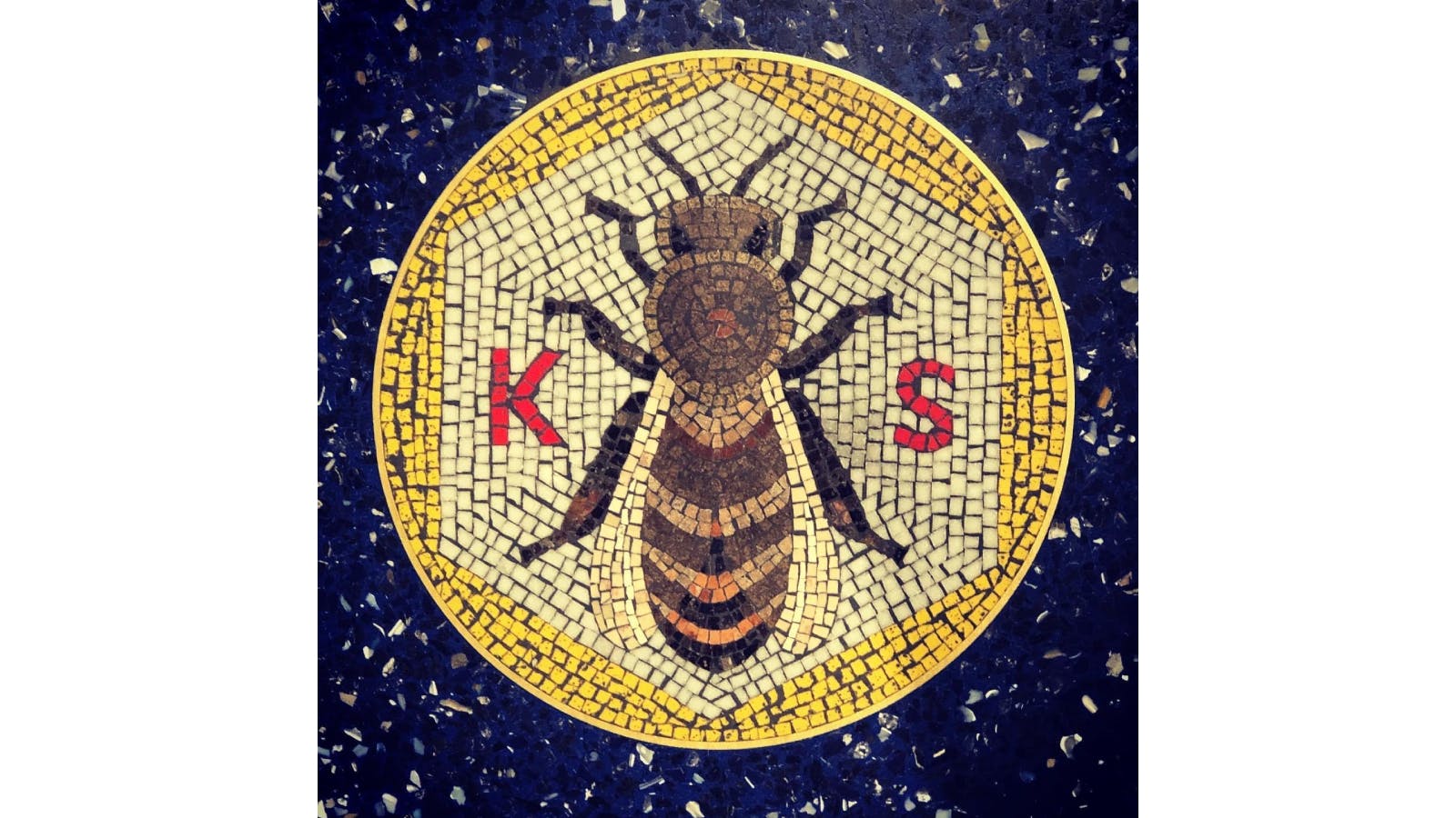 Mosaic of a bee inside a yellow circle on a blue speckled floor at Kansas City International Airport, with red letters "K" and "S" on either side.