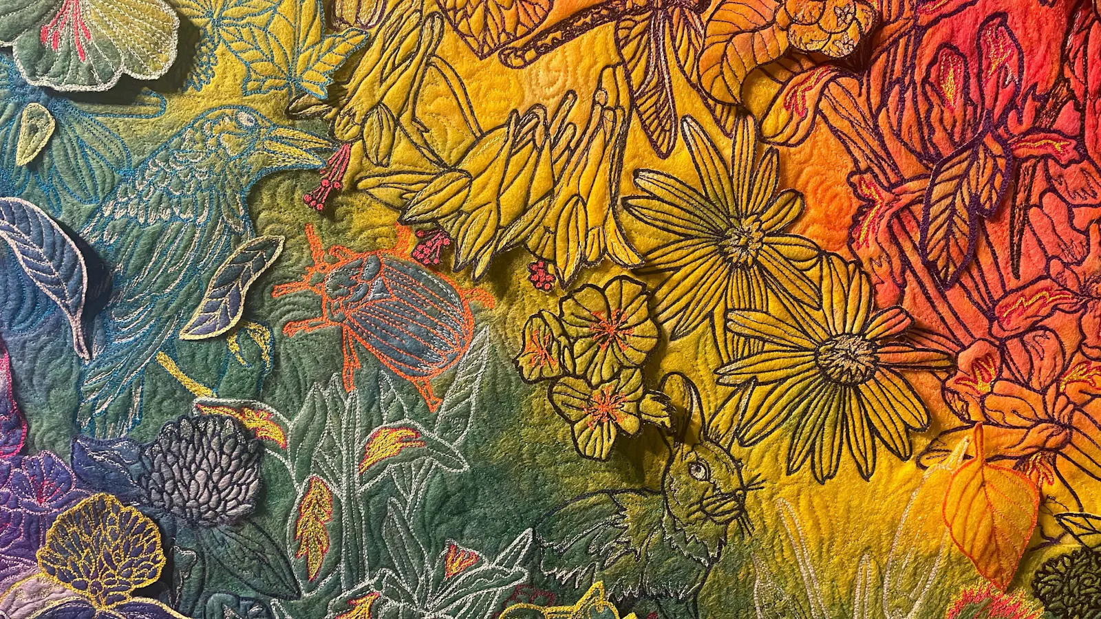 Close-up of rainbow-hued embroidered fabric featuring detailed flowers, leaves, a bird, beetles, dragonflies, and a rabbit stitched into vibrant yellow, green, and red threadwork.