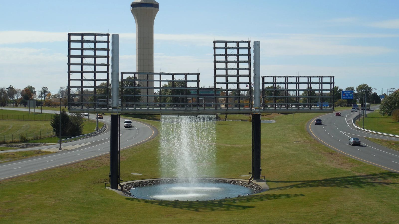 Large fountain structure with water falling from an elevated frame, set between airport roadways with the air traffic control tower in the background.