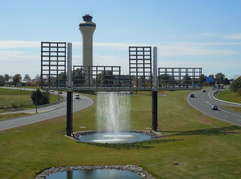 Large fountain structure with water falling from an elevated frame, set between airport roadways with the air traffic control tower in the background.