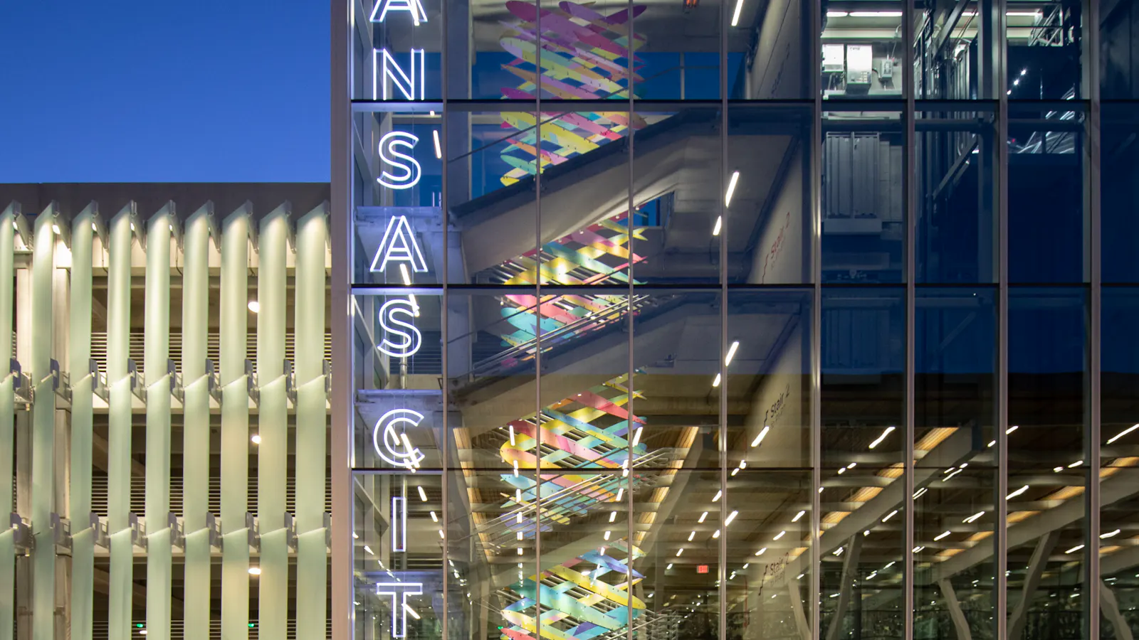 Exterior view of Kansas City International Airport parking garage at dusk, showing a glass stairwell with colorful art inside and a vertical neon sign reading “Kansas City.”