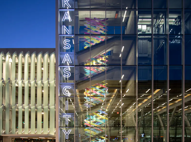 Exterior view of Kansas City International Airport parking garage at dusk, showing a glass stairwell with colorful art inside and a vertical neon sign reading “Kansas City.”